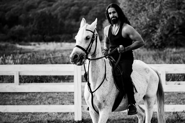 秋の牧場で白い馬に座っている魅力的な男。田舎の彼の種馬に座っている若いハンサムな男の全長。自然に乗って彼の乗馬の男の乗馬。
