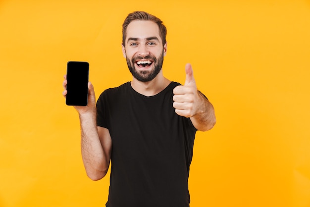 attractive man wearing basic black t-shirt showing thumb up and holding cellphone isolated over yellow wall