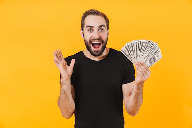 attractive man wearing basic black t-shirt smiling and holding money cash isolated over yellow wall