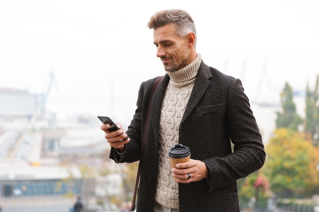 Attractive man wearing jacket walking outdoors, holding cup of takeaway coffee, using mobile phone