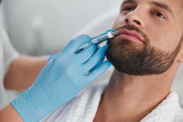 Attractive man with beard undergoes lip augmentation procedure with cosmetologist in clinic