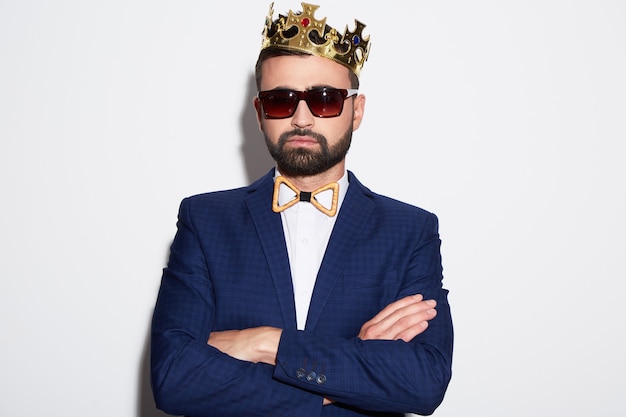 Photo attractive man with black hair and beard wearing white shirt, suit and sunglasses at white studio background, portrait, posing, wearing crown.