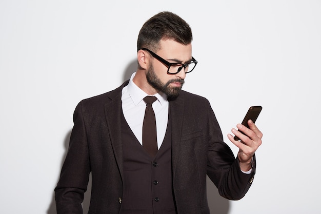 Attractive man with black hair and beard wearing white shirt with tie, black suit and sunglasses at white studio background, portrait, looking at mobile phone.