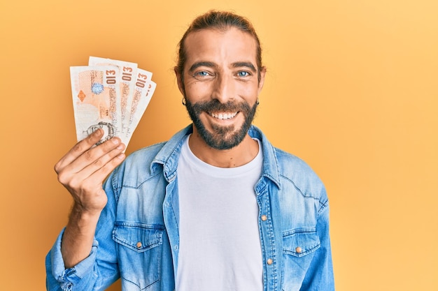 Attractive man with long hair and beard holding 10 united kingdom pounds banknotes looking positive and happy standing and smiling with a confident smile showing teeth