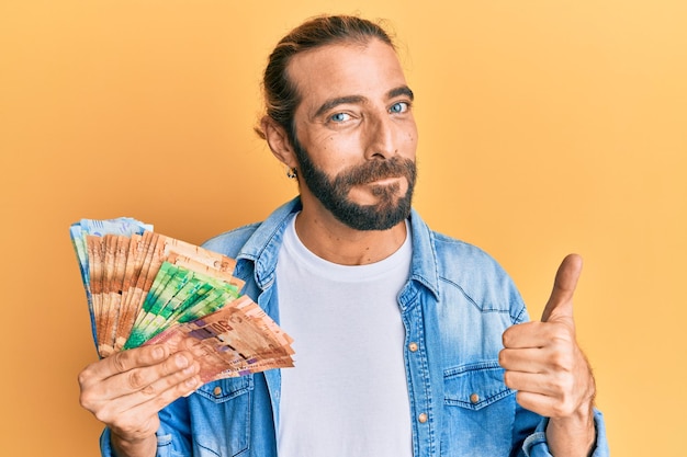 Привлекательный мужчина с длинными волосами и бородой, держащий банкноты южноафриканского ранда, улыбающийся счастливый и позитивный большой палец вверх, делающий отлично и знак одобрения
