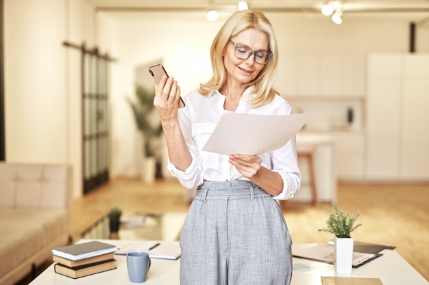 Attractive mature blonde businesswoman having call on speakerphone discussing papers standing in