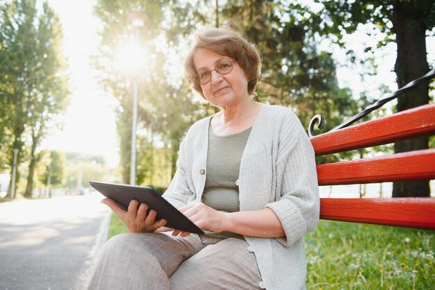 Attractive mature woman sitting on a bench holding and using a digital tablet in the park at summer day