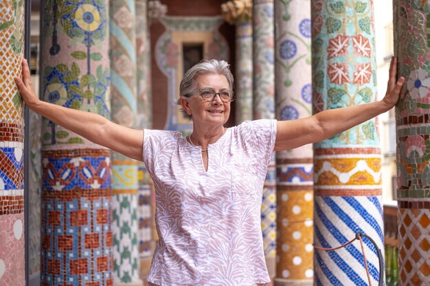 バルセロナの歴史的建造物のカラフルなモザイクの柱の間に立っている魅力的な成熟した女性。休暇を楽しんでいる両手を広げて笑顔の女性