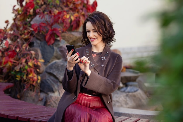 都市公園のベンチに座っている間スマートフォンを使用して魅力的なミドルアデの女。秋の時間。オンライン電話で作業して美しい笑顔の女性