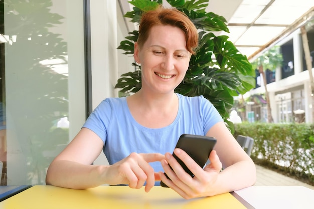 Attractive middle age woman sitting at a table in street cafe smiles while looking at the phone