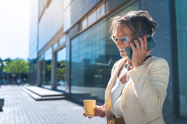 コーヒー カップ通りと彼女のスマート フォンで話している笑顔の魅力的な中年ビジネス女性