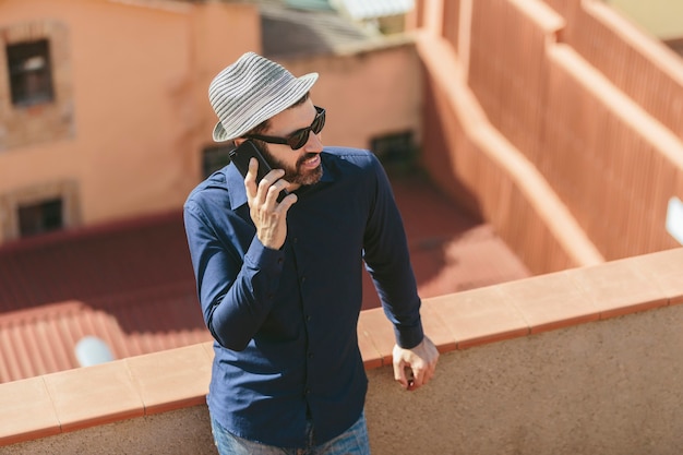 Attractive middle-aged man with hat and sunglasses talking to a smart phone standing on balcony.