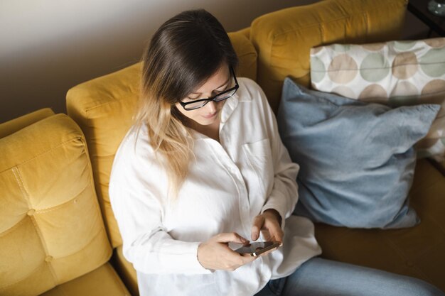 Привлекательная брюнетка средних лет в очках с помощью мобильного телефона сидит на желтом диване в