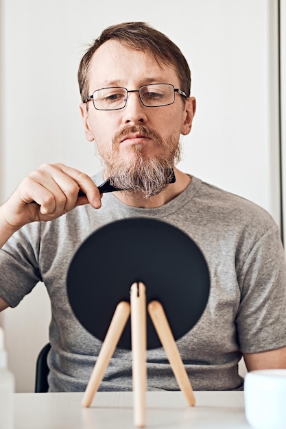 Привлекательный мужчина средних лет поправляет бороду перед зеркалом Естественный вид светло-русые волосы и борода с веснушками и седыми волосами Мягкий фокус