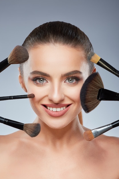 Attractive model with brown hair fixed behind, clean fresh skin, big eyes and naked shoulders posing at gray studio background, close up, portrait, using make up brushes and smiling.