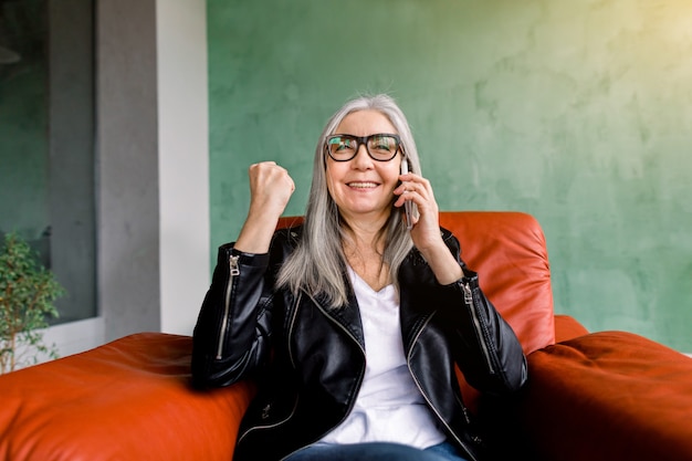 Attractive modern senior lady in black leather jacket, sitting in red soft armchair, keeping her fist clenched and smiling during mobile call