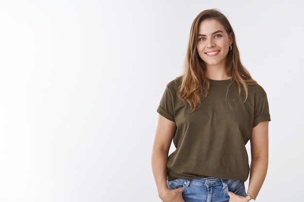 Attraente e moderna giovane donna urbana castagna taglio di capelli corto che indossa t-shirt verde oliva tenendosi per mano tasche sorridente amichevole rilassata posa sana di mente, donna in uscita che comunica sogghignando