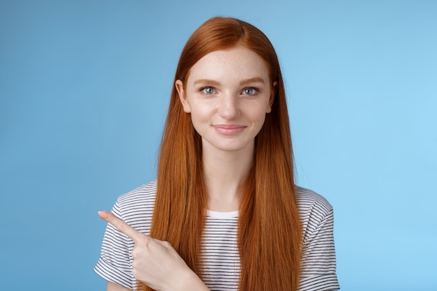 Attraente modesta bella ragazza rossa che agisce maturo amichevole sorridente che punta il dito indice sinistro che dà direzione mostra modo bagno in piedi sfondo blu gioioso sorriso gentile.