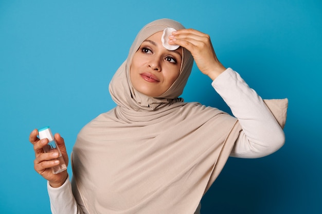 Attraente donna musulmana rimuovere il trucco dal viso utilizzando acqua micellare e batuffolo di cotone.