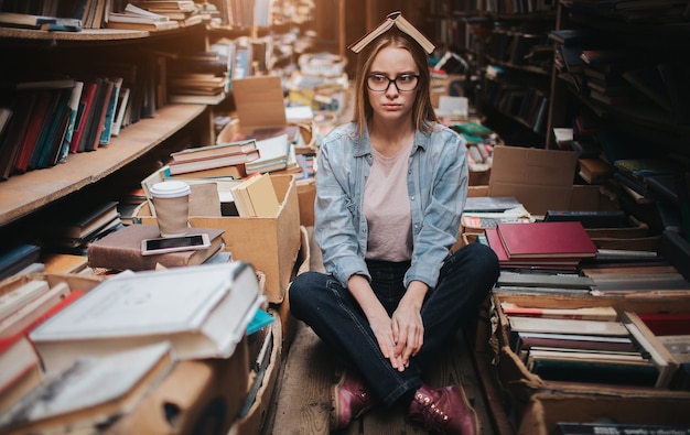 Attractive and nice girl is standing among big and long bookshelfs with old books. She is holding a cup of coffe in her hand and looking to the shelf. The woman is looking for a book