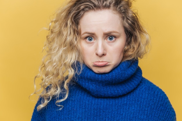 Attractive offended young female has curly bushy hair curves\
lower lip in dissatisfaction feels upset after quarrel with husband\
wears blue sweater isolated over yellow studio background