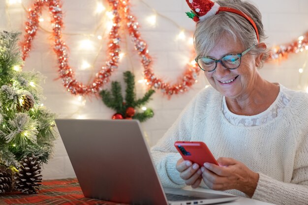 Attraente vecchia donna anziana che utilizza il telefono cellulare sorridente indossando il cappello di babbo natale decorazioni natalizie