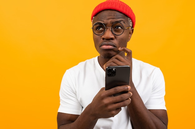 Attractive pensive charming black man in a white T-shirt with a smartphone in his hands on a yellow studio background 