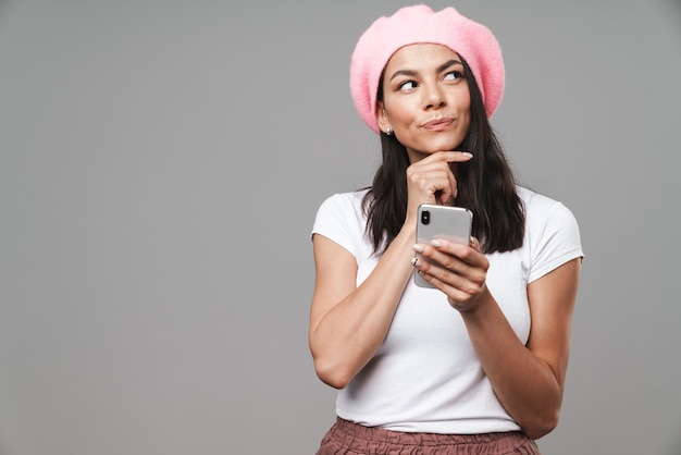 Attractive pensive young brunette woman wearing beret standing isolated over gray wall, holding mobile phone