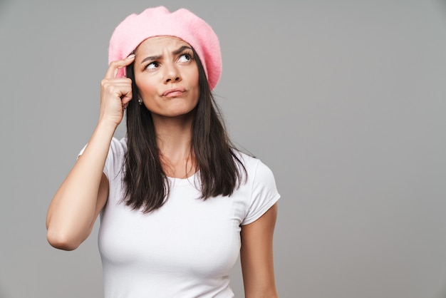 Attractive pensive young brunette woman wearing beret standing isolated over gray wall