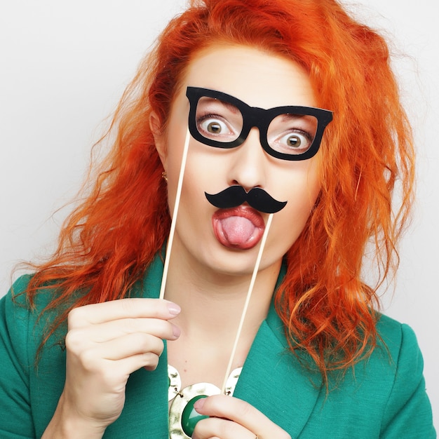 Attractive playful young woman holding mustache and glasses on a stick. Ready for party.