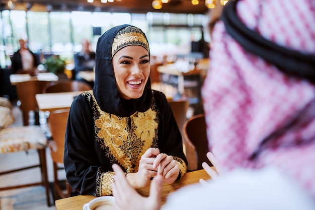 Attractive positive smiling arab woman sitting in cafe with her beloved husband and chatting.