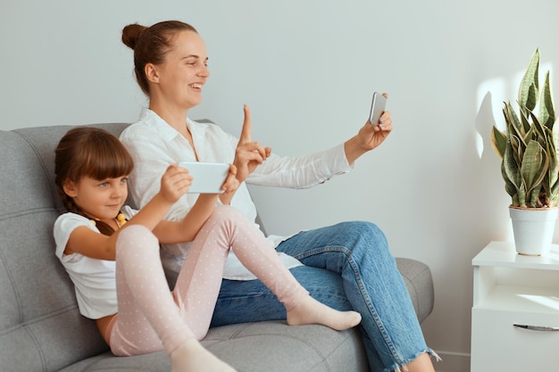 Attractive positive woman with dark hair wearing white shirt and jeans sitting with her daughter on cough and holding phone and showing v sign to device camera, having livestream or video call.