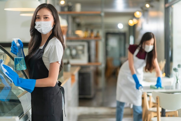 制服のエプロンを着た魅力的なポジティブな若いアジア人女性スタッフが、朝のカフェを安全かつ清潔に消毒するときに、カウンター製品のキャビネットに洗剤またはアルコールをスプレーして柔らかい布で拭きます