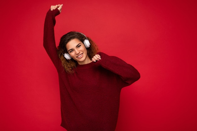 Attractive positive young brunet curly woman wearing dark red sweater isolated over red background