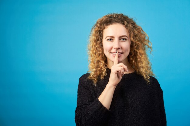 Attractive positive young redhead curly woman keeps finger at her mouth, asks to keep a secret.