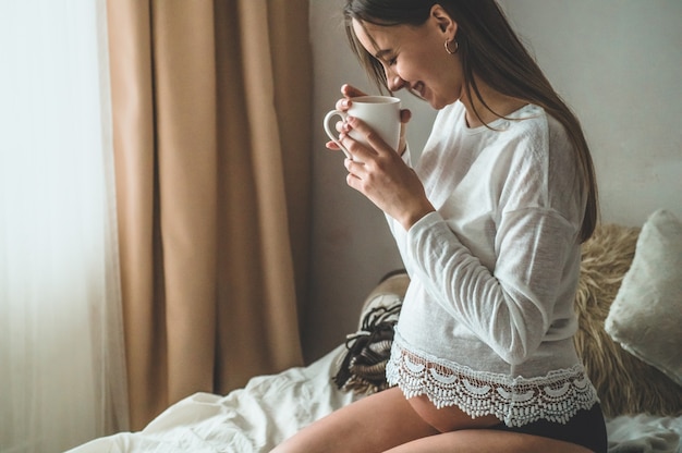 Attractive pregnant woman drinks tea on the bed. Drinking tea looking through a window at home. Last months of pregnancy.