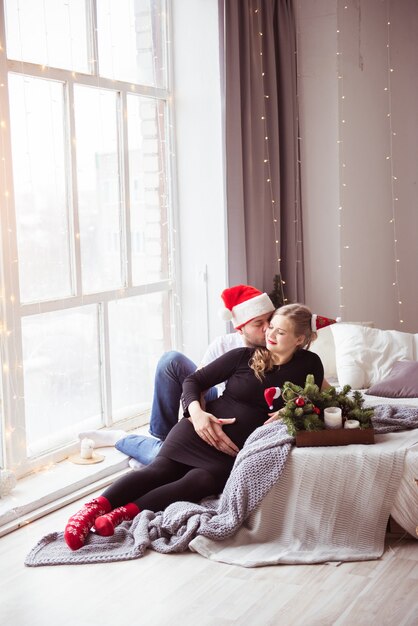 Una donna incinta attraente e un bell'uomo in un abbraccio di cappello da babbo natale su un letto bianco