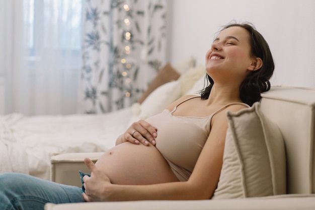 魅力的な妊婦は肘掛け椅子に座って腹を抱えています。