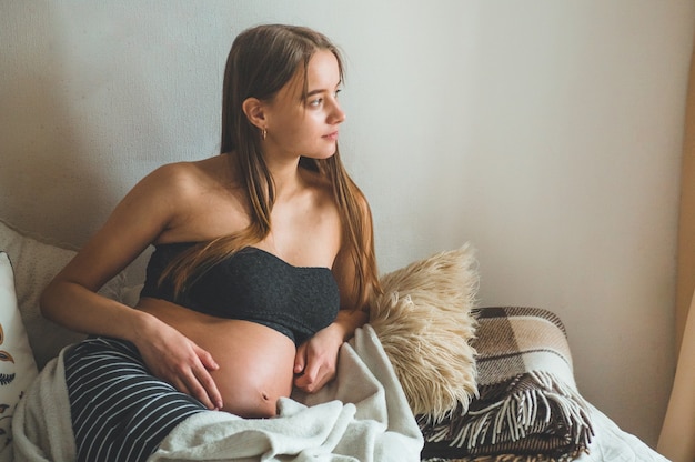 Attractive pregnant woman is sitting in bed and holding her belly. Last months of pregnancy.