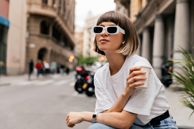 Foto attraente bella signora con i capelli corti in occhiali da sole che indossa abiti casual trascorre del tempo all'aperto con un caffè nella mattina d'estate graziosa signora che tiene una tazza di tè in posa durante il riposo all'aperto