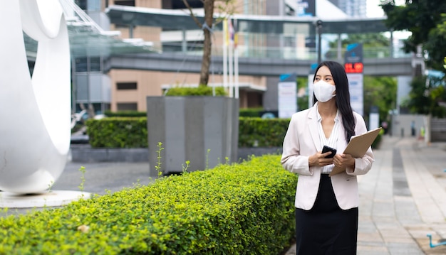 보호용 얼굴 마스크를 쓴 휴대전화를 사용하는 매력적인 아시아 비즈니스 여성은 야외에서 코비드-19 바이러스를 예방합니다