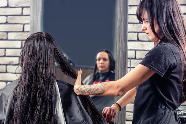 Attractive professional hairdresser combing wet hair of young brunette woman while she is sitting in armchair in beauty salon