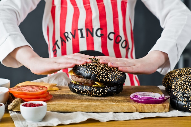 An attractive red-haired boy in a chef's apron is cooking a hamburger in the kitchen. Recipe for cooking black cheeseberger. Homemade juicy Burger.