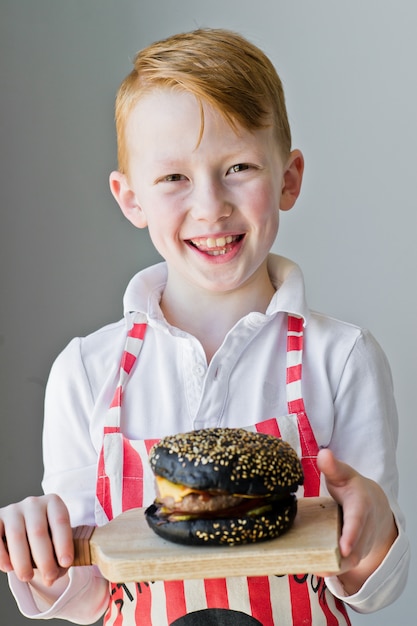 An attractive red-haired boy is holding a wooden chopping Board with a hamburger.