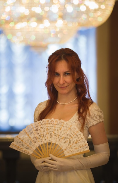 Attractive red haired woman in vintage  centuries dresses use fan in luxury ballroom