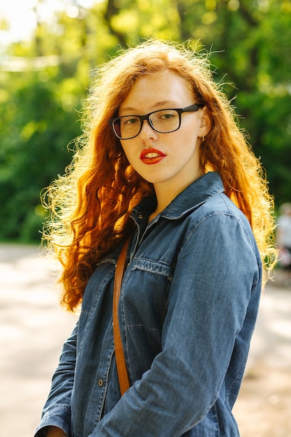 Attractive red haired young woman in jeans shirt and glasses posing at the street