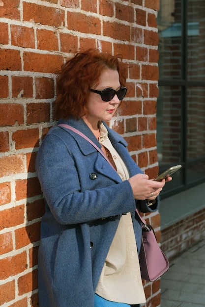 Привлекательная рыжеволосая женщина в синем пальто держит смартфон