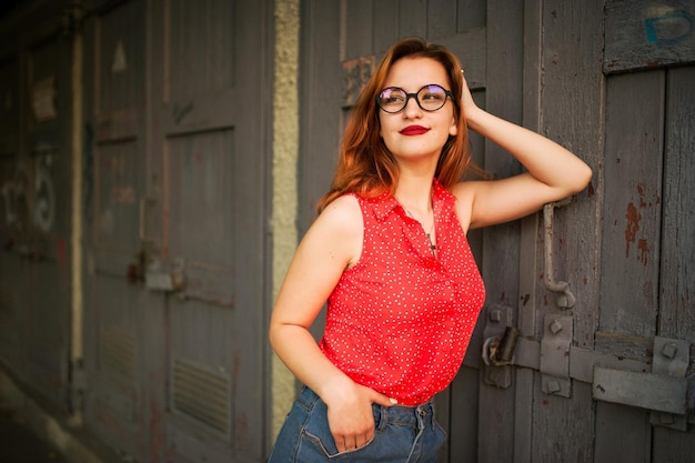 Attractive redhaired woman in eyeglasses wear on red blouse and jeans skirt posing