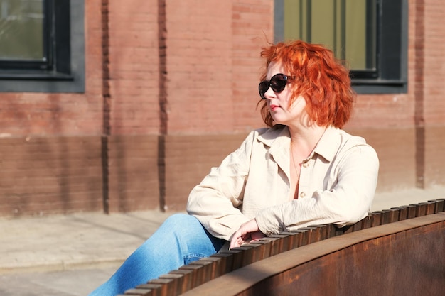 Attractive redhaired woman with sunglasses sits on a bench in a business district