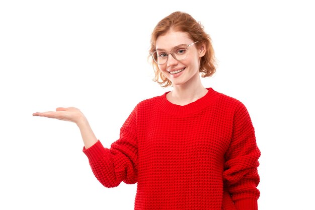 Attractive redhead girl promoter in red sweater holding something in hand demonstrating empty space with excited face isolated on white background